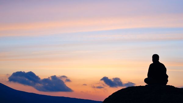The shadow of a person sitting on a hill, overlooking mountains and a sunset with clouds of purple and coral.